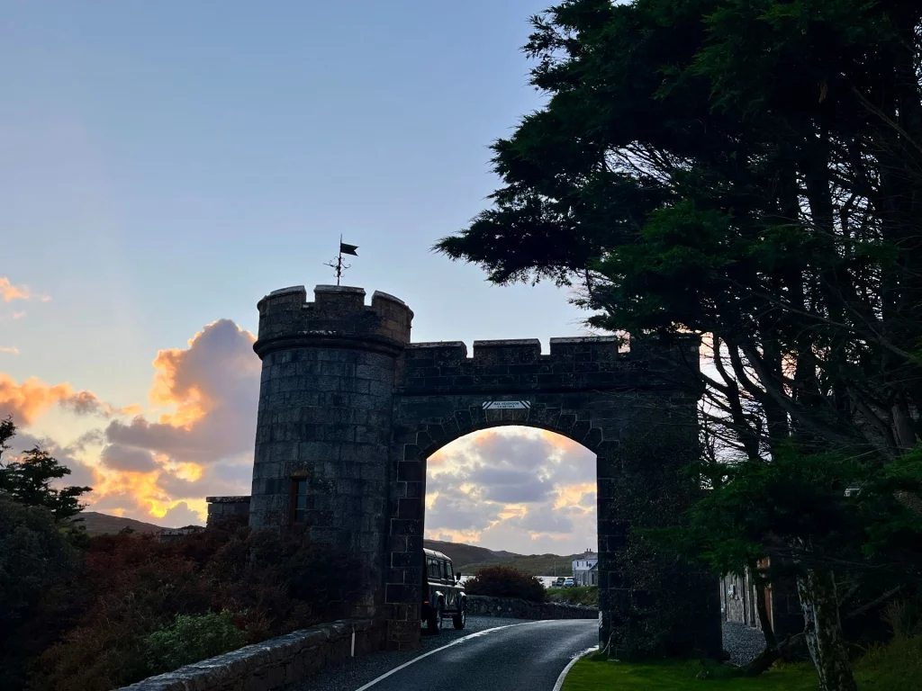 Ein massives, burgähnliches Tor aus dunklem Stein mit einem runden Zinnenturm auf der linken Seite. Die Straße führt unter dem Torbogen hindurch in Richtung einer Küstenlandschaft. Im Hintergrund leuchten die Wolken im goldenen Licht des Sonnenuntergangs, während im Vordergrund dunkle Silhouetten von Bäumen und Gebüsch den Rahmen bilden.