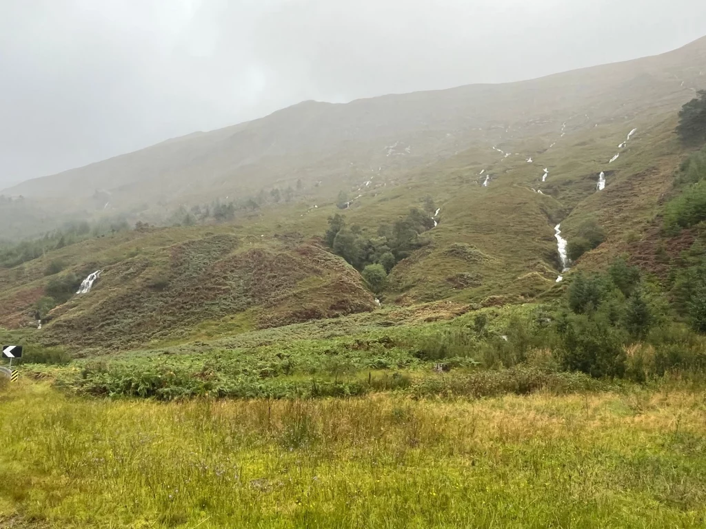 Blick auf einen weiten, nebligen Hang in einer kargen Berglandschaft. Mehrere kleine Wasserfälle ziehen sich wie weiße Adern durch das braun-grüne Grasland.
