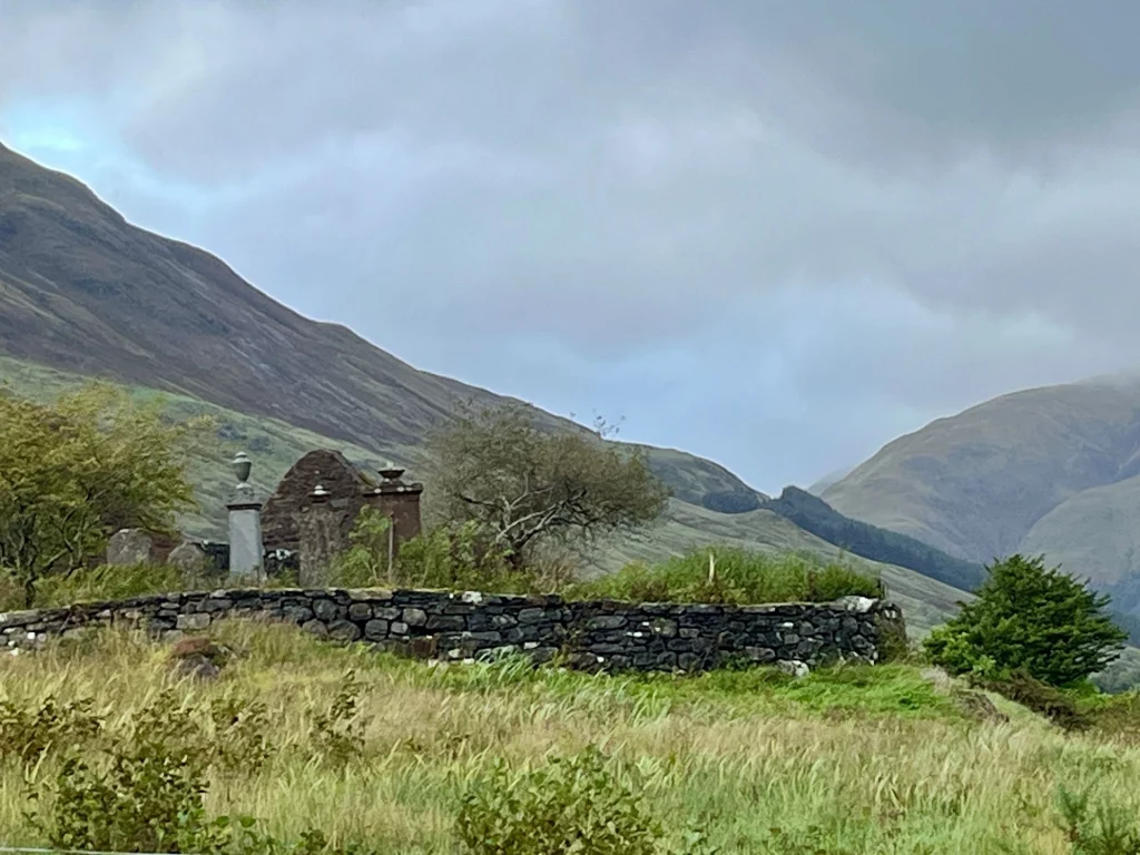 Eine Nahaufnahme einer alten Trockensteinmauer und der Überreste eines Steingebäudes vor einem grünen Berghang. Im Vordergrund wiegt sich hohes, helles Gras im Wind unter einem bedeckten Himmel.