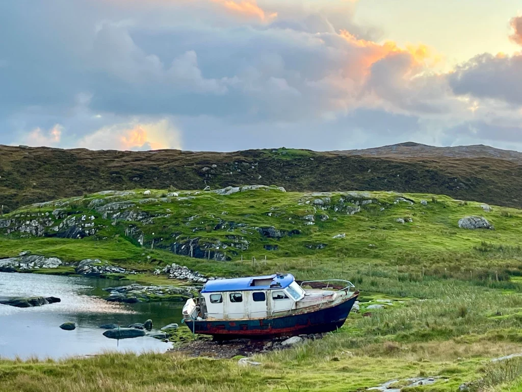 Ein altes, blau-weißes Fischerboot liegt verlassen am grasbewachsenen Ufer einer Bucht, im Hintergrund erheben sich sanfte grüne Hügel unter einem dämmerigen Abendhimmel.
