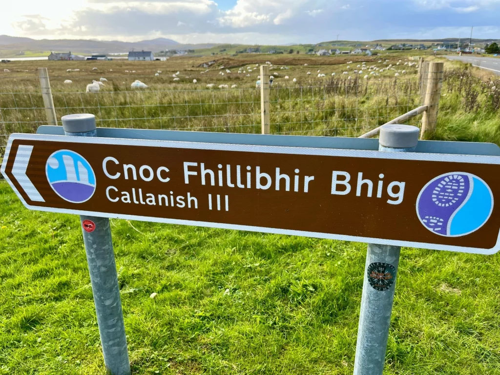 Ein braunes Hinweisschild aus Metall mit der Aufschrift 'Cnoc Fhillibhir Bhig / Callanish III' weist den Weg zu den historischen Steinkreisen in einer ländlichen Umgebung.