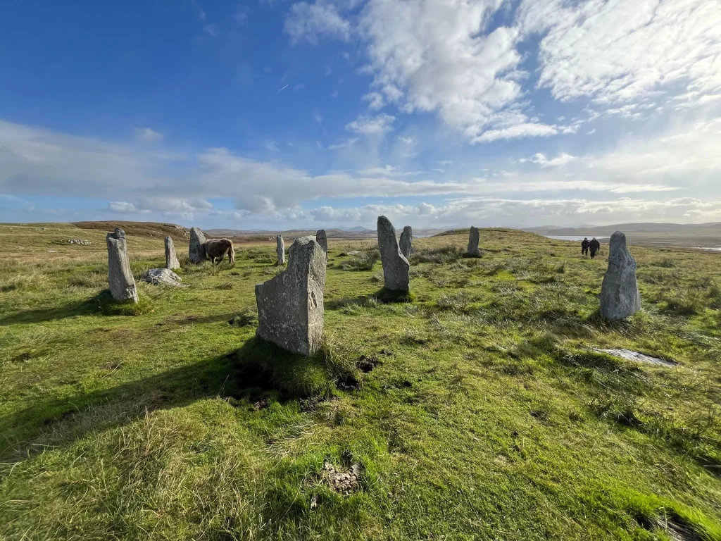 Weitwinkelaufnahme eines antiken Steinkreises auf einer grünen, hügeligen Wiese unter einem weiten, leicht bewölkten blauen Himmel.