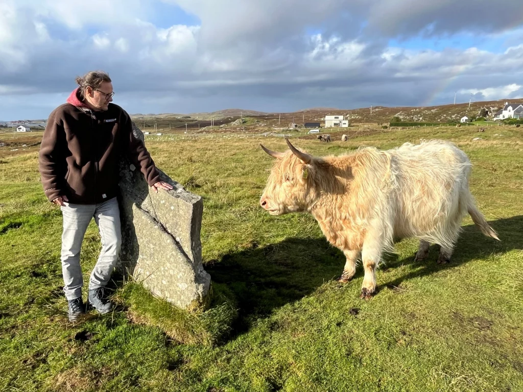 Ingo, in seiner braunen Jacke, steht an einem der Megalithen und blickt zu einem hellbeigen Hochlandrind hinüber, das friedlich auf der sonnigen Wiese steht.
