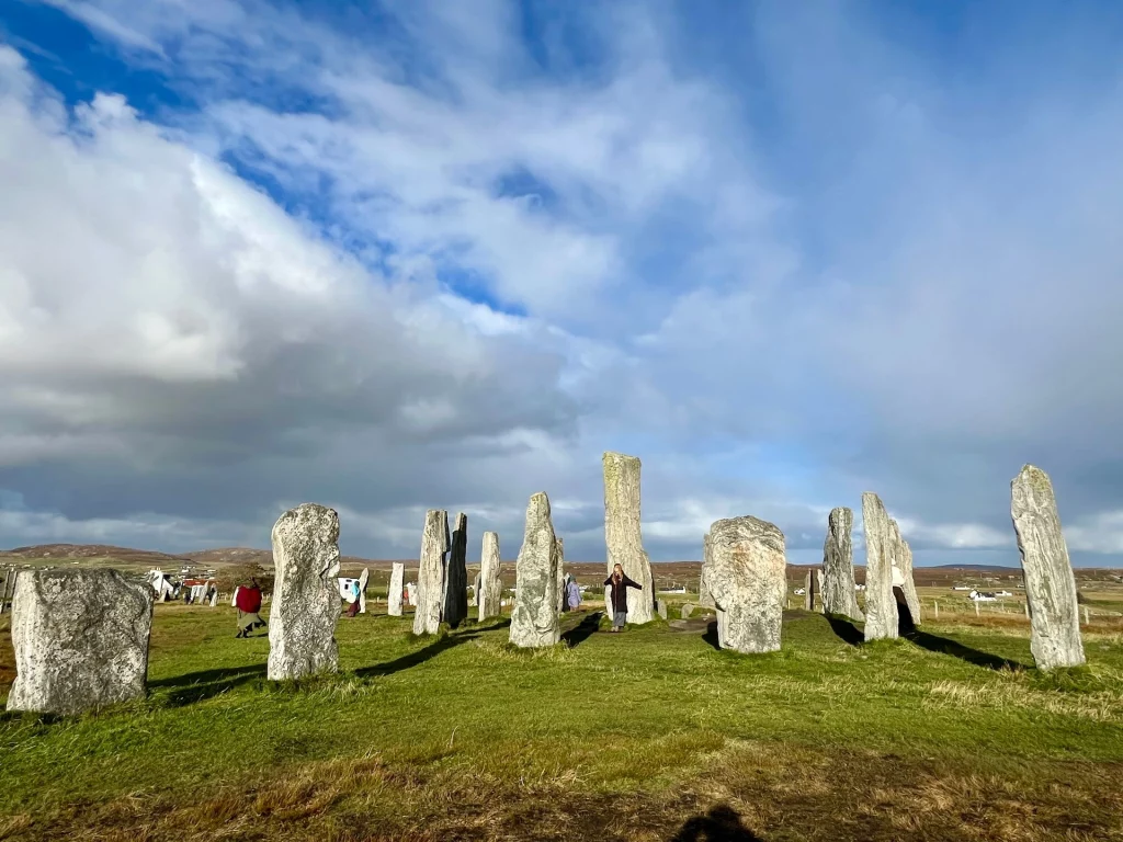 Zahlreiche aufrecht stehende Megalithen des Callanish-Steinkreises auf einer grünen Wiese unter einem dramatischen Wolkenhimmel, durch den die Sonne hell und kreisförmig hindurchscheint.