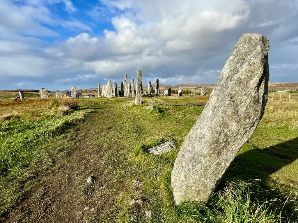 Blick an einem schrägen Monolithen vorbei auf den fernen Steinkreis von Callanish bei hellem Tageslicht.