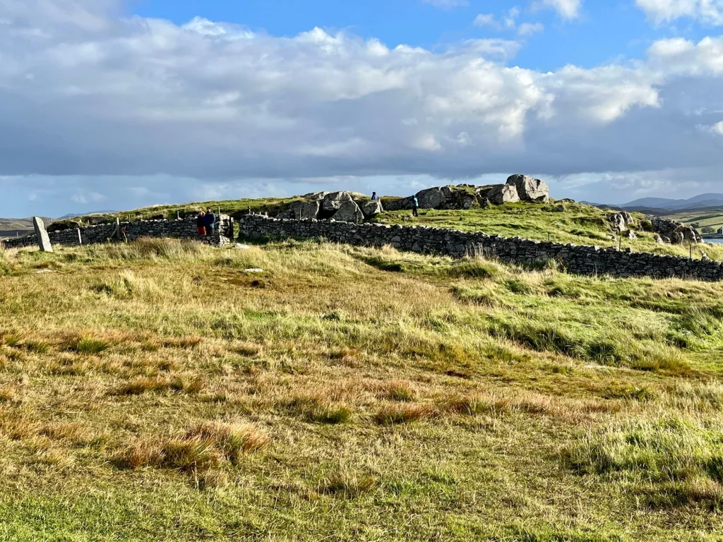Weitwinkelansicht des Steinkreises von Callanish auf einem grasbewachsenen Hügel unter blauem Himmel mit weißen Wolken, begrenzt durch eine traditionelle Steinmauer.