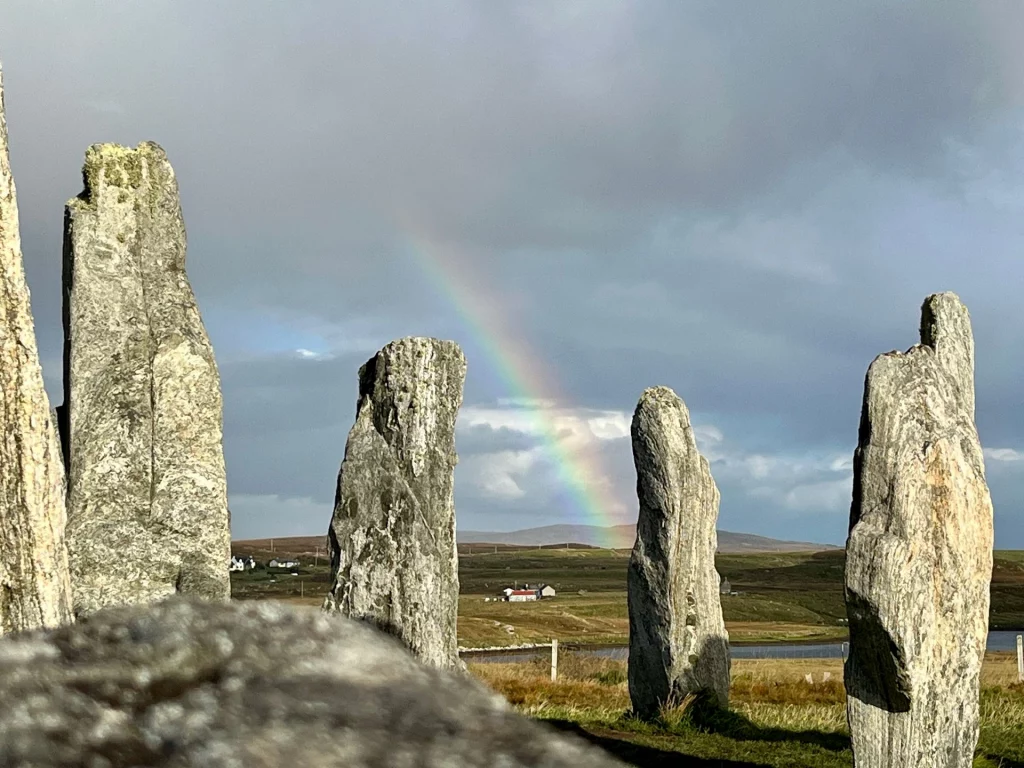 Hohe, graue Monolithen des Callanish-Steinkreises im Vordergrund mit einem leuchtenden Regenbogen über der kargen schottischen Hügellandschaft im Hintergrund.