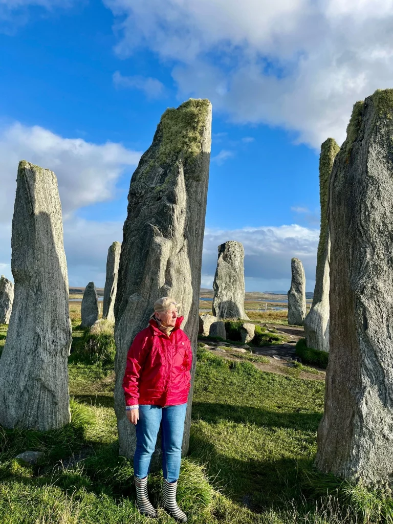 Eine Frau in einer roten Regenjacke und blauen Jeans steht zwischen den hohen, grauen Steinstelen von Callanish. Die Steine sind teilweise mit gelblichem Moos bewachsen. Der Himmel ist strahlend blau mit weißen Wolken, und das Sonnenlicht wirft lange Schatten auf das grüne Gras der historischen Anlage.