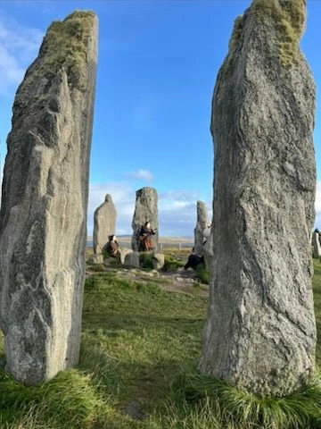 Eine Perspektivaufnahme durch zwei massive, hohe Steinpfeiler hindurch auf das Zentrum des Steinkreises. Im Hintergrund sind weitere Steine und die weite, flache Hügellandschaft der Isle of Lewis unter einem klaren blauen Himmel zu sehen. Die Textur des uralten Gesteins im Vordergrund ist detailreich erkennbar.
