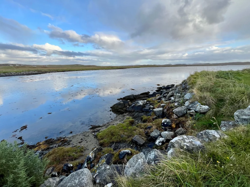 Eine weite Naturaufnahme einer Bucht oder eines Sees bei klarem Wetter. Das Ufer im Vordergrund ist mit moosbewachsenen Steinen und grünem Gras gesäumt. Die Wasseroberfläche spiegelt den blau-weißen Wolkenhimmel perfekt wider, während am Horizont sanfte, flache Hügelketten verlaufen.