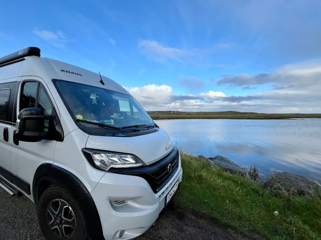 Ein hellgrauer Knaus Kastenwagen-Camper steht auf einem befestigten Weg direkt an einem ruhigen See oder Meeresarm. Der Himmel ist blau mit vereinzelten Wolken, die sich im glatten Wasser spiegeln. Im Hintergrund ist eine weite, flache Heidelandschaft unter spätnachmittäglichem Licht zu sehen.