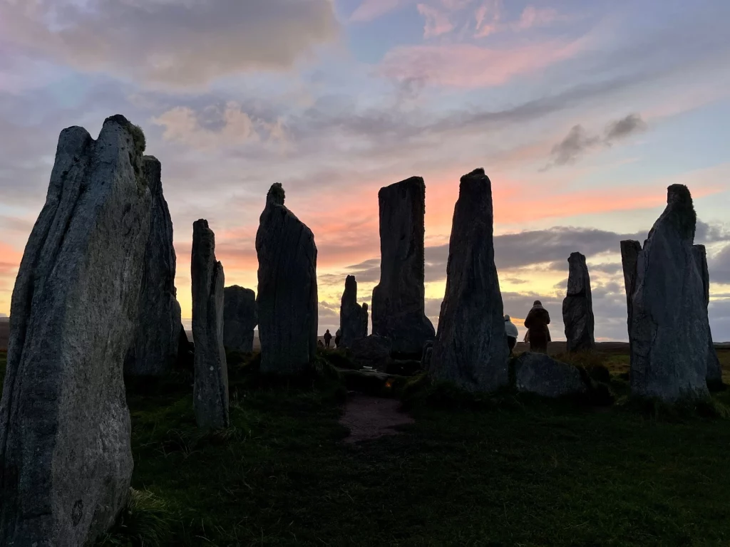 Die Steine von Callanish als dunkle Silhouetten vor einem dramatischen Sonnenuntergang in Orange- und Rosatönen. Einzelne Gestalten verlieren sich fast zwischen den riesigen Monolithen.