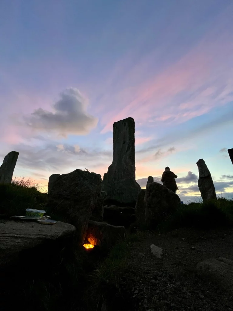 Eine dramatische Silhouette der Callanish Stones gegen einen violett-rosa gefärbten Abendhimmel. Im Vordergrund ist eine dunkle Steinstruktur zu sehen, in deren Vertiefung ein warmes Licht (wie eine Kerze oder kleine Lampe) leuchtet. In der Ferne sind die Umrisse einer weiteren Person zwischen den Steinen erkennbar.