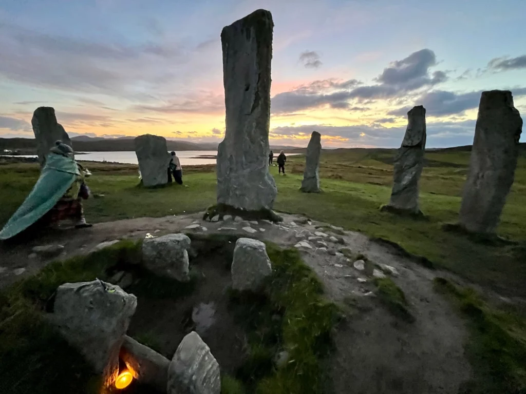 Eine Weitwinkelaufnahme der Anlage in der Dämmerung. Im Vordergrund brennt ein kleines Licht in einer Vertiefung, während links eine Person in eine grüne Decke gehüllt an einem der Steine vorbeigeht. Im Hintergrund glitzert das Wasser eines fernen Lochs.