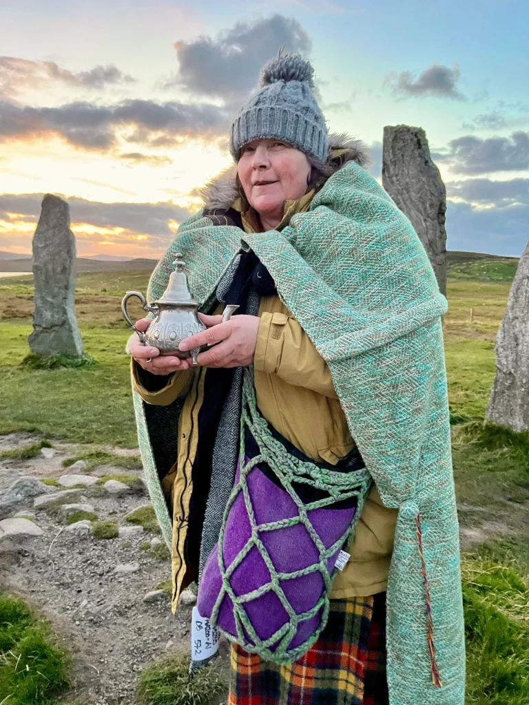 Eine Frau steht lächelnd vor den Callanish Standing Stones auf der Isle of Lewis während eines farbenprächtigen Sonnenuntergangs. Sie trägt eine graue Strickmütze, eine gelbe Outdoorjacke und ist in eine große, grün-türkis gewebte Decke gehüllt. In ihren Händen hält sie eine kleine, verzierte silberne Teekanne. Über ihrer Schulter hängt eine auffällige violette Tasche mit grobem Netzmuster, und im unteren Bildrand ist ein traditioneller Schottenkaro-Rock (Kilt) zu sehen. Im Hintergrund ragen die historischen Megalith-Steine in den dämmernden Himmel.