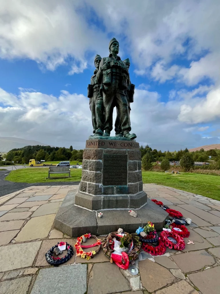 Eine Bronzestatue von drei Commando-Soldaten des Zweiten Weltkriegs auf einem hohen Steinsockel vor einem weiten, bewölkten Himmel in Schottland. Am Fuß des Denkmals liegen zahlreiche Gedenkkränze und Blumen. Auf dem Sockel steht die Inschrift „United We Conquer“.