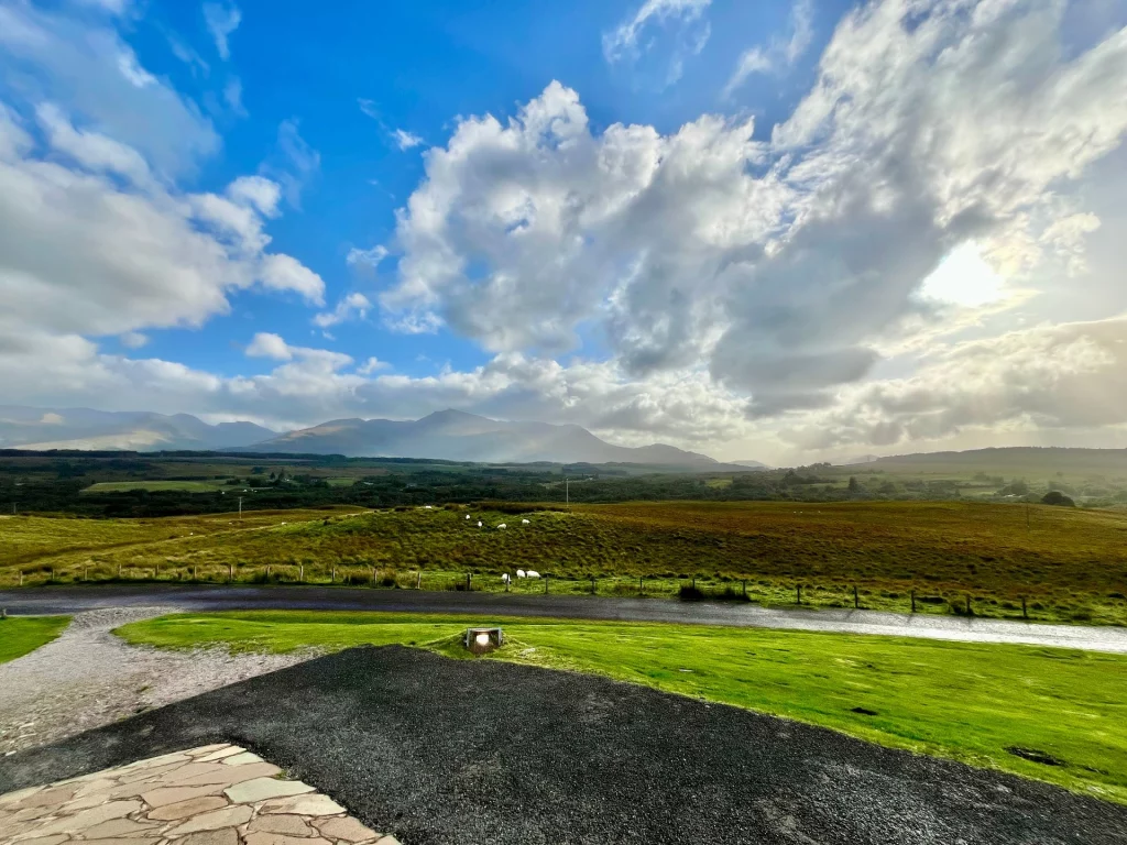 Eine weitläufige schottische Hügellandschaft unter einem weiten blauen Himmel. Eine massive, hell erleuchtete Wolkenformation steht im Zentrum, hinter der die Sonne hervorstrahlt und Lichtstrahlen auf die grünen Felder wirft. Im Hintergrund sind dunkle Bergsilhouetten zu sehen.