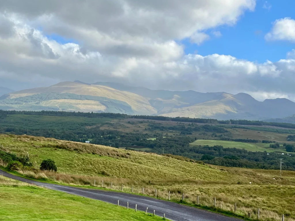 Weitwinkelaufnahme einer grünen Hügellandschaft in Schottland. Im Hintergrund erstreckt sich die Bergkette des Ben Nevis unter einem dynamischen Wolkenhimmel. Im Mittelgrund grasen einige weiße Schafe auf einer Weide, die von einem einfachen Zaun begrenzt wird.