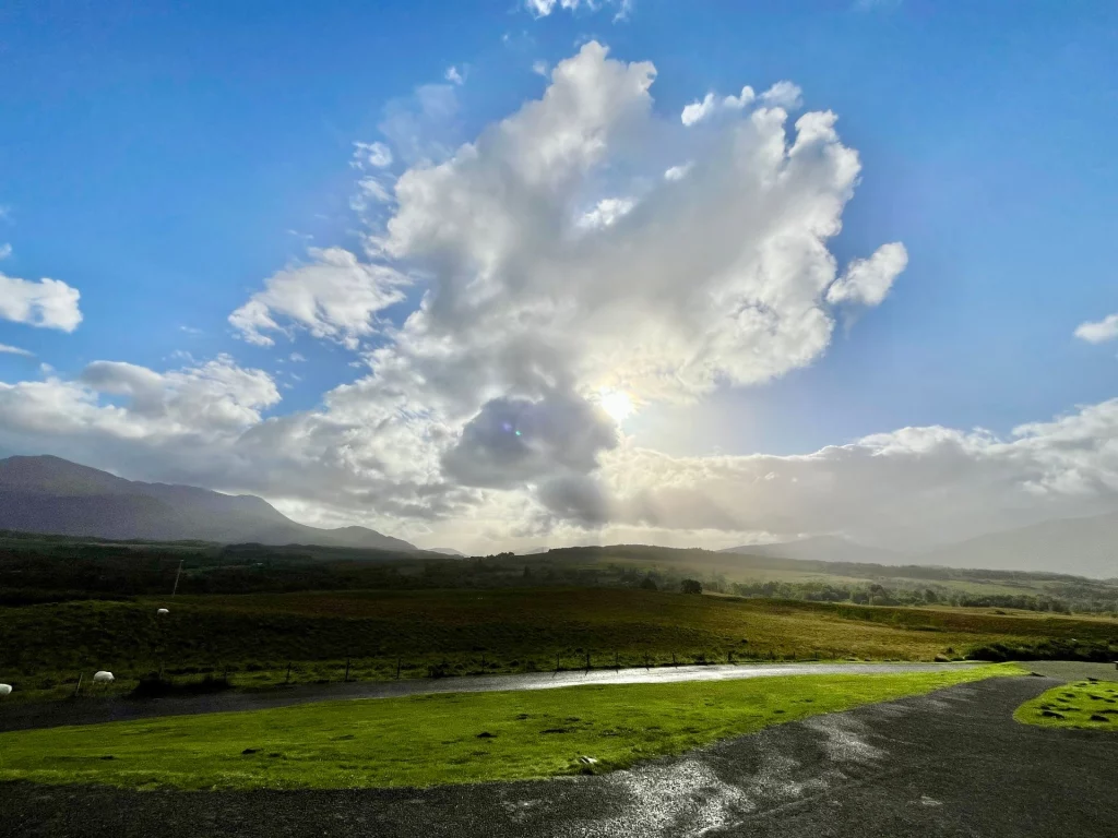 Eine Detailaufnahme der schottischen Hochlandlandschaft. Sanfte, teils bewaldete Hügel steigen zu den massiven, grasbewachsenen Flanken der Berge an. Eine schmale Asphaltstraße schneidet durch das satte Grün im Vordergrund.