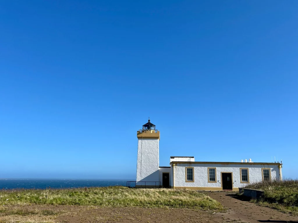 Ein quadratischer, weißer Leuchtturm mit gelben Akzenten und einer schwarzen Laterne steht neben einem einstöckigen weißen Funktionsgebäude. Das Ensemble befindet sich auf einer grasbewachsenen Klippe vor einem tiefblauen Meer und einem wolkenlosen Himmel in Duncasby Head, Schottland.