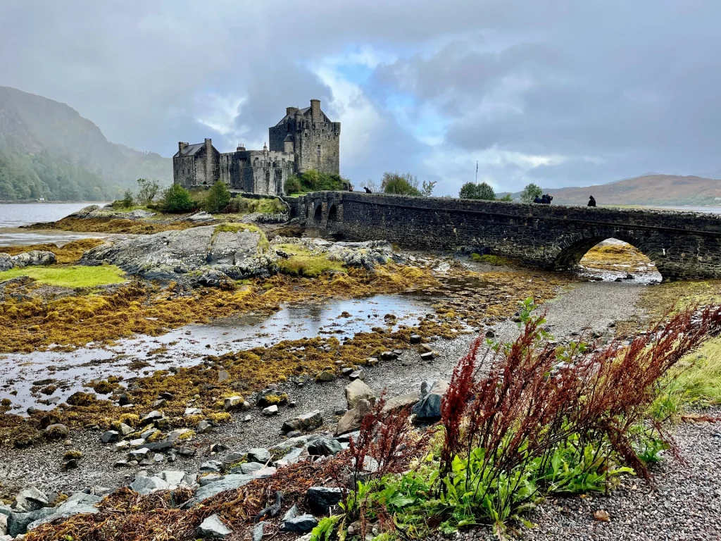 Das Eilean Donan Castle auf seiner kleinen Insel, umgeben von weiten, mit gelb-braunem Seegras bedeckten Flächen bei Ebbe. Im Hintergrund ragen dunkle Berge unter schweren Wolken auf.