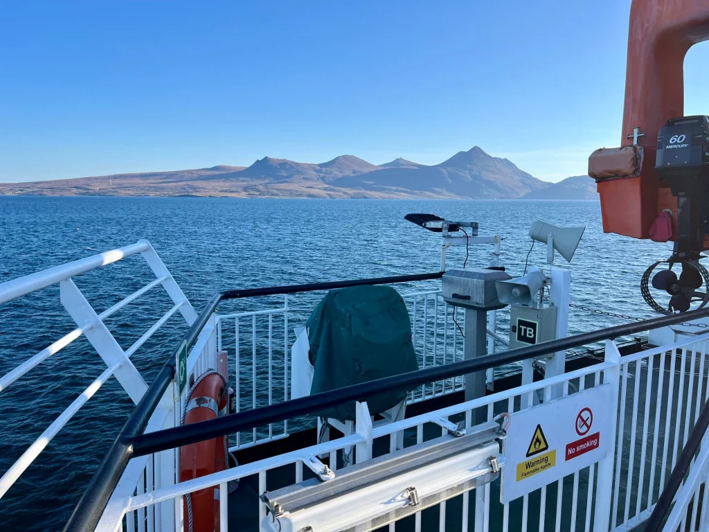 Blick vom weißen Reling-Deck der Fähre über das tiefblaue Meer auf die fernen, bergigen Silhouetten der schottischen Küste.