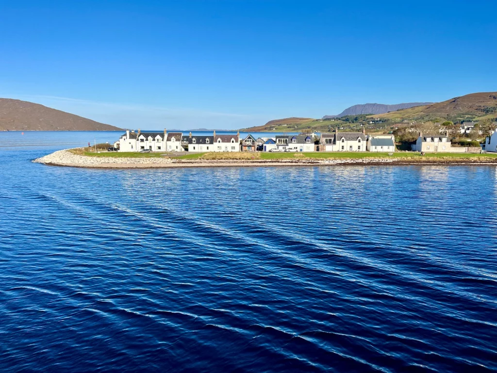 Blick von der Fähre auf die weißen Häuserfronten von Ullapool am Ufer des Loch Broom unter strahlend blauem Himmel in den schottischen Highlands.