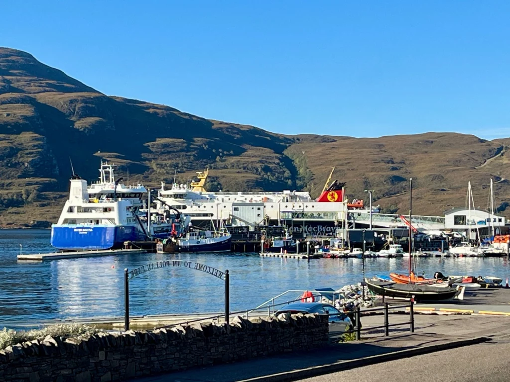 Die große blau-weiße CalMac-Fähre liegt im sonnigen Hafen von Ullapool vor einer gewaltigen Bergkulisse am Kai.
