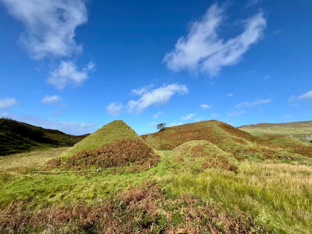 Ein einzelner, perfekt geformter Laubbaum thront einsam auf der Spitze eines runden, grünen Kegelhügels unter blauem Himmel.