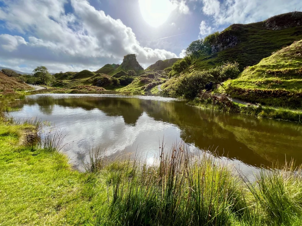 Weitwinkelaufnahme eines stillen, dunklen Teichs im Fairy Glen, in dessen glatter Oberfläche sich die grünen Hügel, der blaue Himmel und die hellen Wolken perfekt spiegeln. Im Hintergrund ragt der Castle Ewen Felsen markant auf, während das Sonnenlicht sanft durch die Wolken bricht.