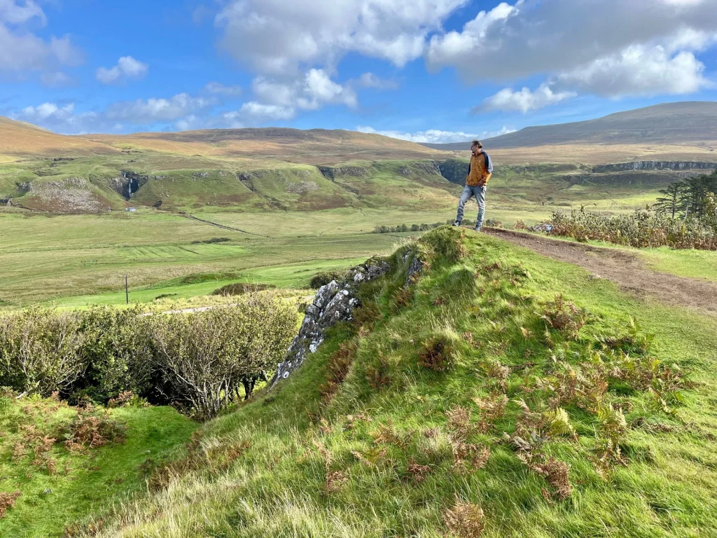 Ein Mann steht triumphierend auf dem schmalen, grasbewachsenen Kamm eines Hügels im Fairy Glen. Im Hintergrund erstreckt sich die weite, sonnige Hügellandschaft von Skye unter einem blauen Himmel.