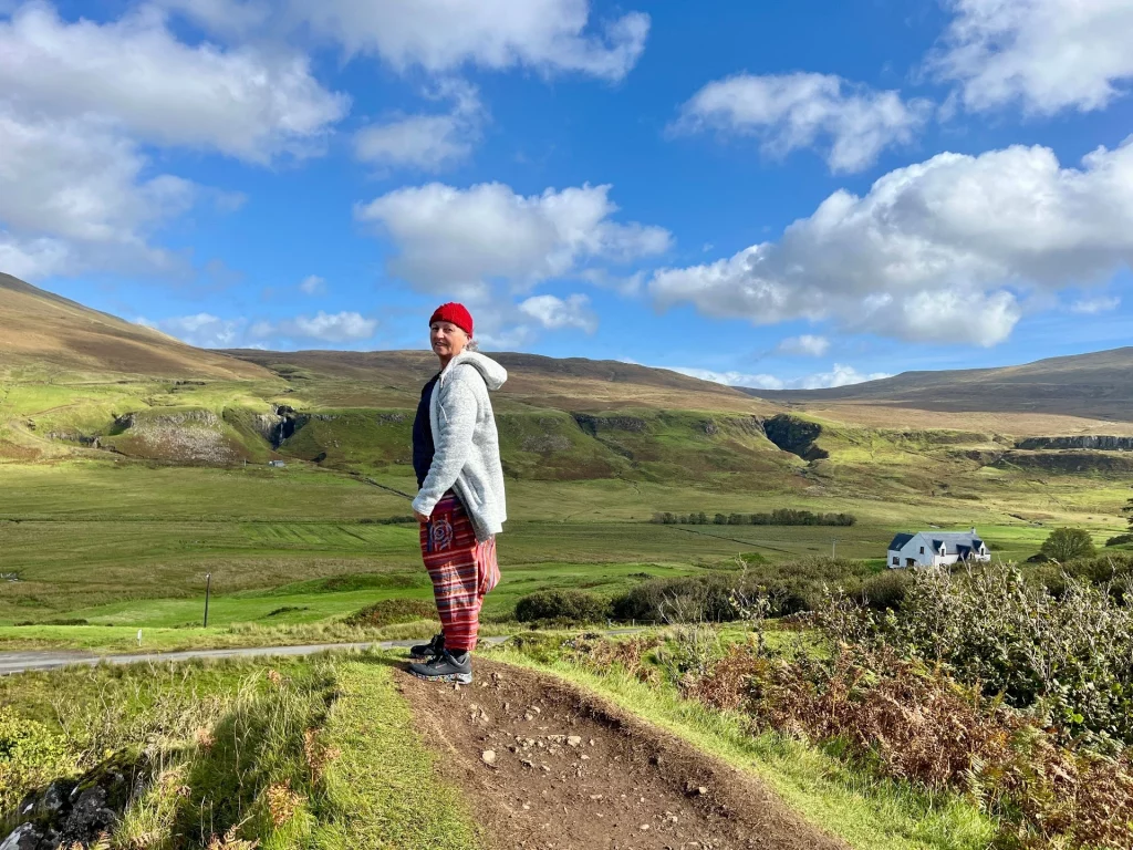 Eine Person mit auffälliger roter Mütze und grauem Mantel steht auf einem erhöhten Pfad und blickt über das weite, grüne Tal. In der Ferne ist ein einsames weißes Haus vor den sanften Bergen zu sehen.