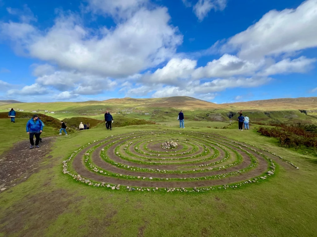 Blick von oben auf eine große, kreisförmige Steinspirale, die von Besuchern im flachen Grasland des Fairy Glen angelegt wurde. In der Mitte der Spirale befindet sich ein kleiner Steinhaufen. Im Hintergrund sind Wanderwege, sanfte grüne Hügel und mehrere Touristen unter einem strahlend blauen Himmel mit dekorativen Schönwetterwolken zu sehen.