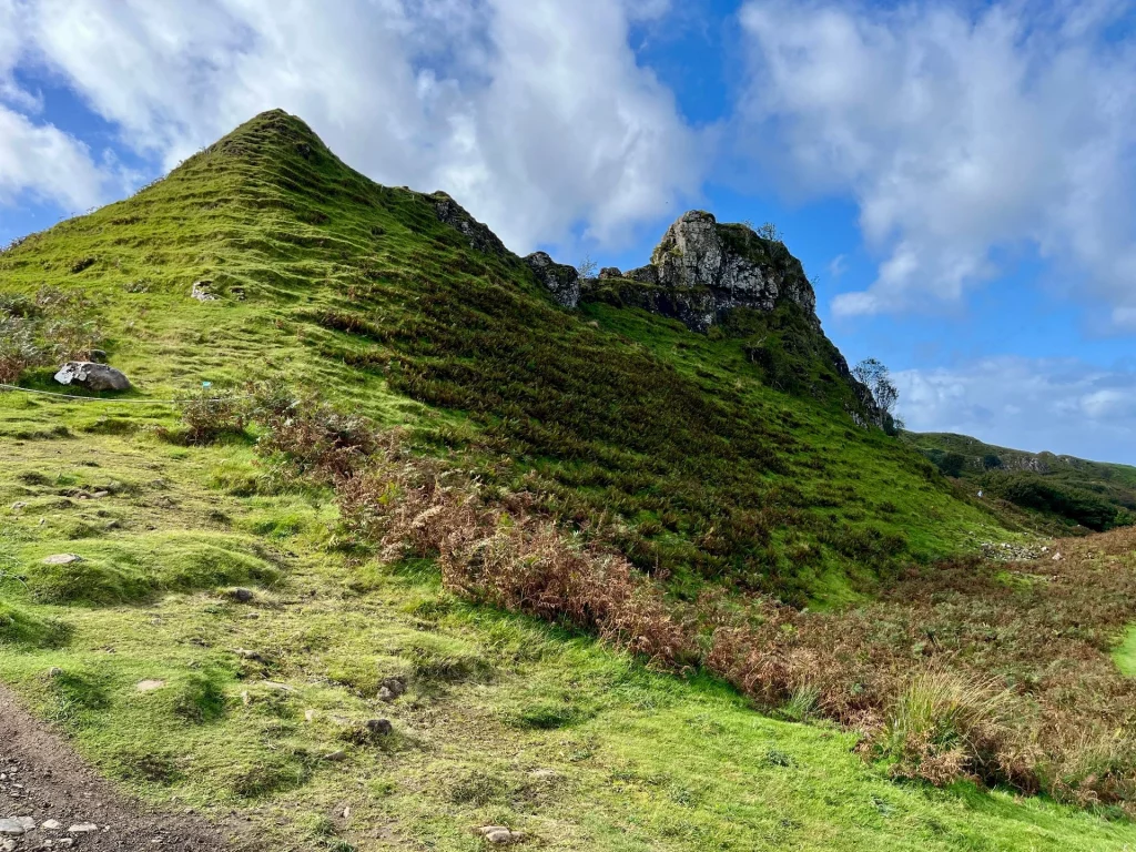 Nahaufnahme eines steilen, kegelförmigen Grashügels im Fairy Glen auf Skye. Der Hügel zeigt horizontale Erosionsspuren (Schafpfade) und ist teilweise mit braunem Farn bewachsen. Im Hintergrund ragt der dunkle Basaltfelsen Castle Ewen in den bewölkten blauen Himmel.