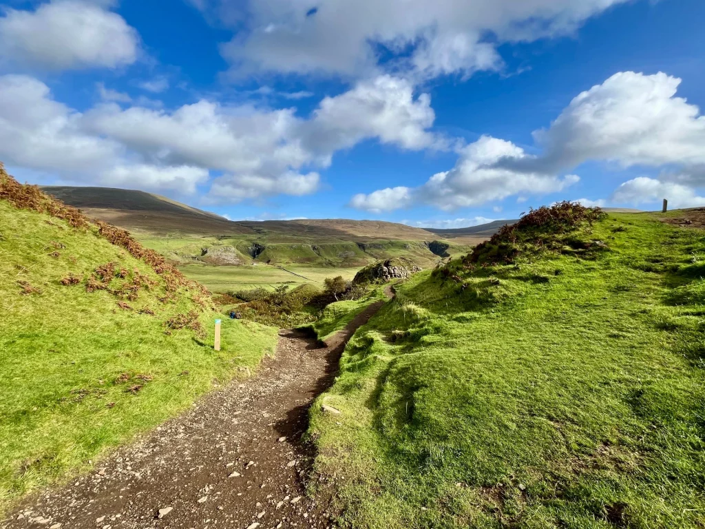 Ein schmaler, erdiger Wanderpfad windet sich zwischen zwei leuchtend grünen Hügeln hindurch. Der Weg führt direkt in die mystische Hügellandschaft des Tals.