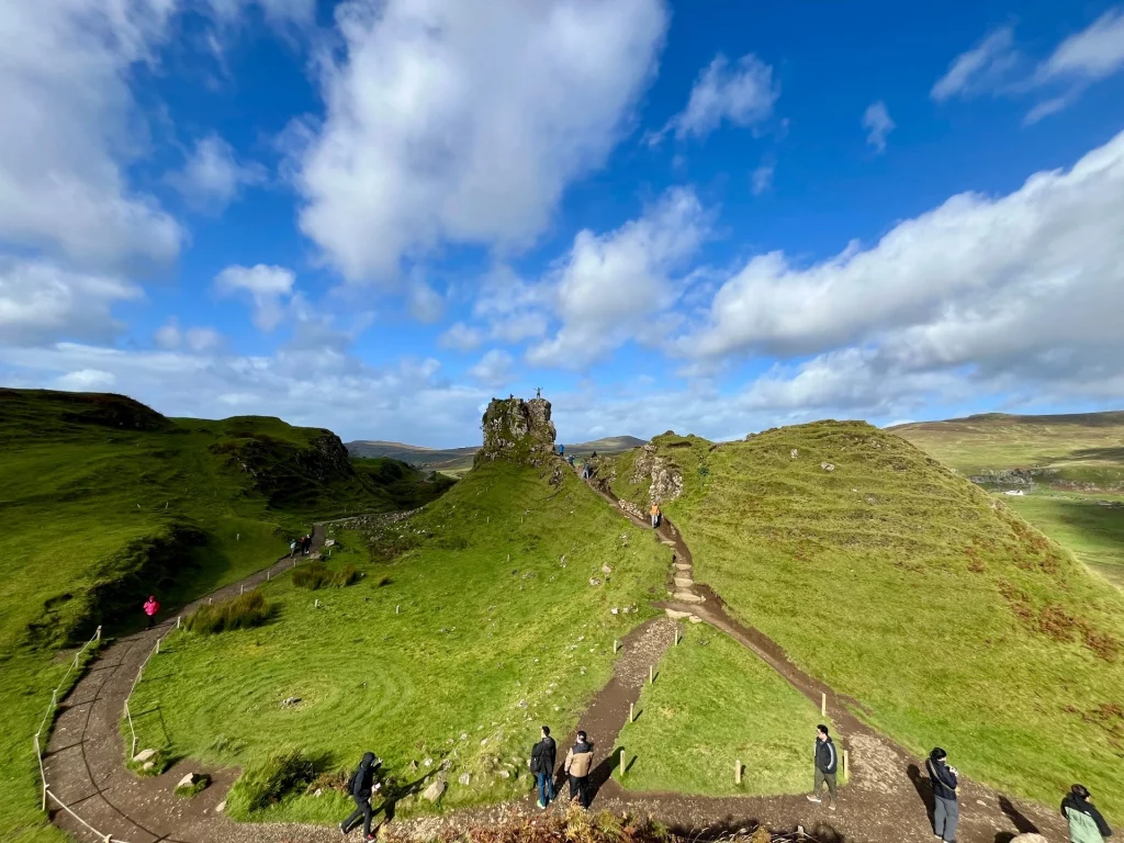Weitwinkelaufnahme des Fairy Glen auf der Isle of Skye unter strahlend blauem Himmel mit weißen Wolken. Man sieht grüne, kegelförmige Hügel, Wanderwege mit mehreren Spaziergängern und das markante Felsplateau „Castle Ewen“ im Zentrum. Im Gras ist links ein spiralförmiges Steinlabyrinth zu erkennen.