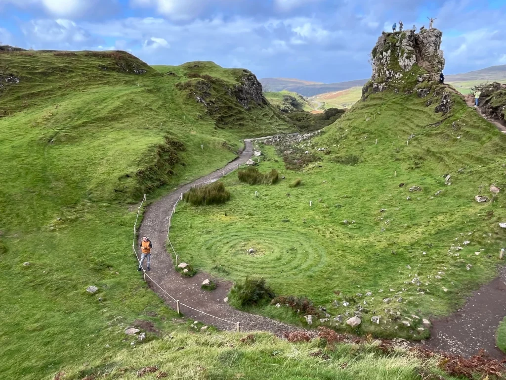 Weitwinkelaufnahme des Fairy Glen auf der Isle of Skye unter strahlend blauem Himmel mit weißen Wolken. Man sieht grüne, kegelförmige Hügel, Wanderwege mit mehreren Spaziergängern und das markante Felsplateau „Castle Ewen“ im Zentrum. Im Gras ist links ein spiralförmiges Steinlabyrinth zu erkennen. 