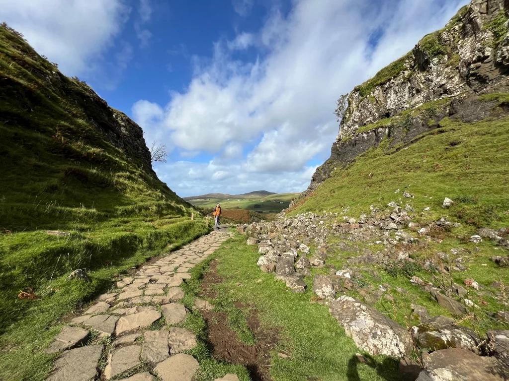 Ein rustikaler Pfad aus unregelmäßigen Steinplatten windet sich durch ein enges, grünes Tal im Fairy Glen. Rechts des Weges liegen lose Felsbrocken am Hang, während im Hintergrund eine Person den Pfad entlangwandert.