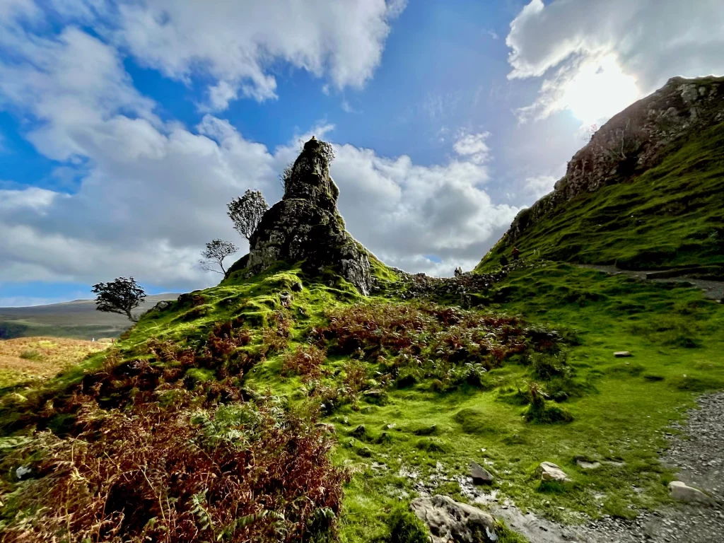 Der markante Castle Ewen Basaltfelsen ragt steil und zerklüftet in den Himmel. Die Sonne steht tief hinter den Wolken und erzeugt ein dramatisches Gegenlicht, das die herbstlich braunen Farne und die grünen Hänge im Vordergrund leuchten lässt.