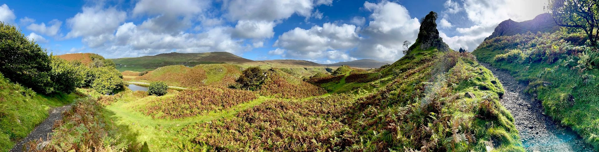 Ein weites Panorama des Fairy Glen auf Skye, das die gesamte surreale Hügellandschaft einfängt. Von dichten, grünen Büschen auf der linken Seite zieht sich der Blick über sanfte, mit braunem Farn bewachsene Wellenformen bis hin zum markanten, spitzen Felszinken des Castle Ewen auf der rechten Seite, flankiert von einem schmalen Wanderpfad unter einem dramatischen Wolkenhimmel.“