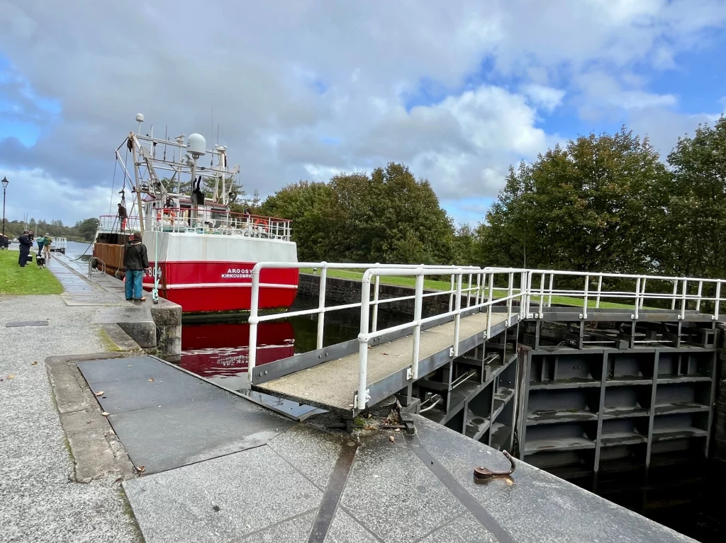 Frontalansicht des rot-weißen Fischkutters „Argosy“ in der engen Steinschleuse des Caledonian Canal in Fort William, Passanten beobachten das Manöver vom Rand aus.