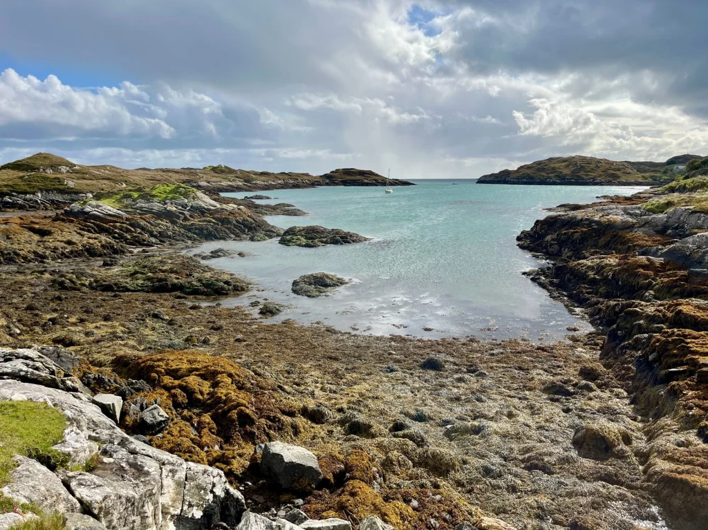 Blick auf eine ruhige Meeresbucht mit klarem, türkisfarbenem Wasser, das von dunklen, flachen Felsen und braunem Seetang eingerahmt ist. Die Küstenlinie ist zerklüftet und grün bewachsen. Über dem Horizont hängen schwere Regenwolken, durch die vereinzelt Licht auf das Wasser fällt.