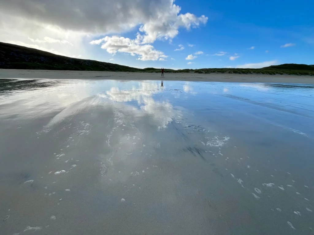 Weitwinkelaufnahme eines nassen Sandstrandes bei Ebbe. Das flache Wasser reflektiert den blauen Himmel und die weißen Wolken so perfekt, dass die Grenze zwischen Boden und Horizont verschwimmt.
