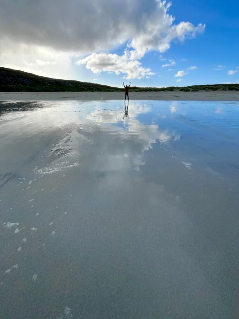 Weitwinkelaufnahme eines einsamen Strandes bei Ebbe. Im Hintergrund steht eine kleine menschliche Silhouette mit triumphierend erhobenen Armen. Der extrem flache, nasse Sand im Vordergrund wirkt wie ein perfekter Spiegel, der den blauen Himmel, die weißen Quellwolken und die Person präzise reflektiert. Flache, grüne Hügel begrenzen den Strandabschnitt unter einem weiten, lichtdurchfluteten Himmel.