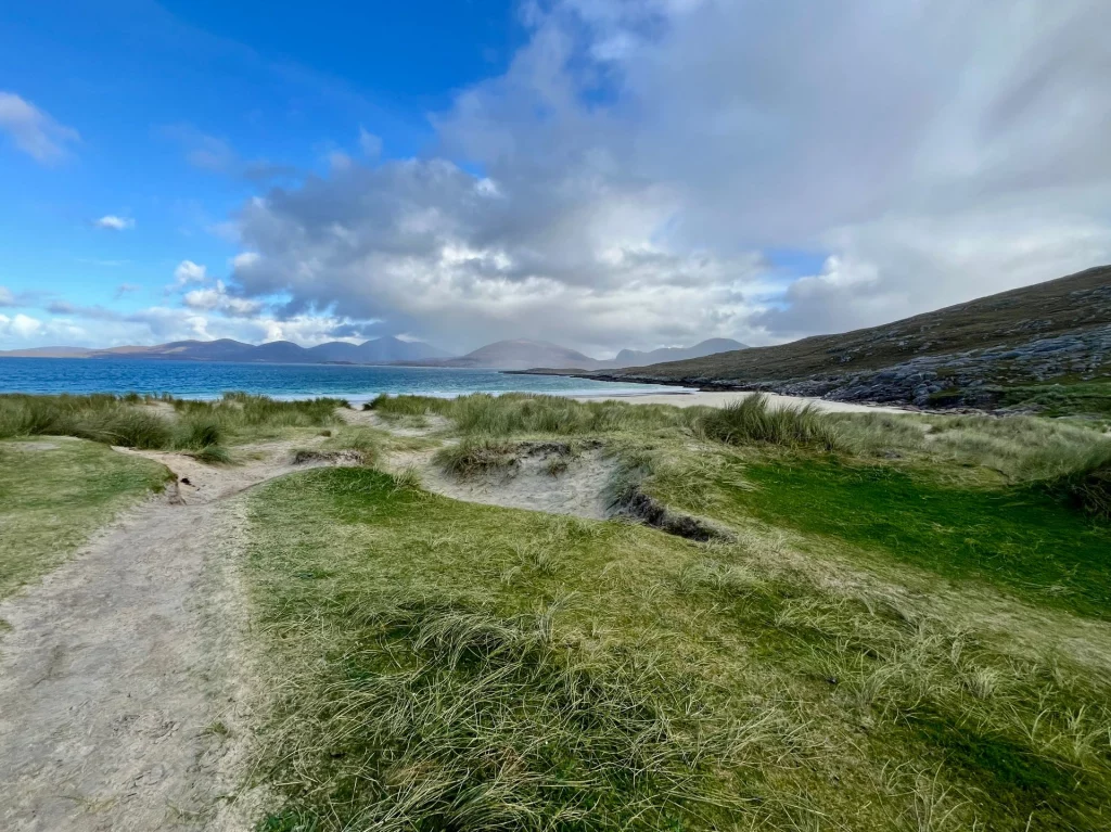 Blick über eine grasbewachsene Dünenlandschaft hinweg auf eine Meeresbucht. Im Hintergrund sind dunkle Hügelketten unter einem wechselhaften Wolkenhimmel zu sehen.