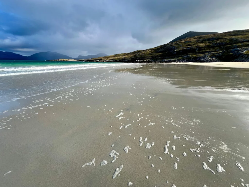 Blick über den nassen Sandstrand auf das türkisblaue Wasser. Weiße Schaumflocken der Brandung liegen verstreut im Vordergrund auf dem dunklen Sand, dahinter ragen dunkle Klippen auf.