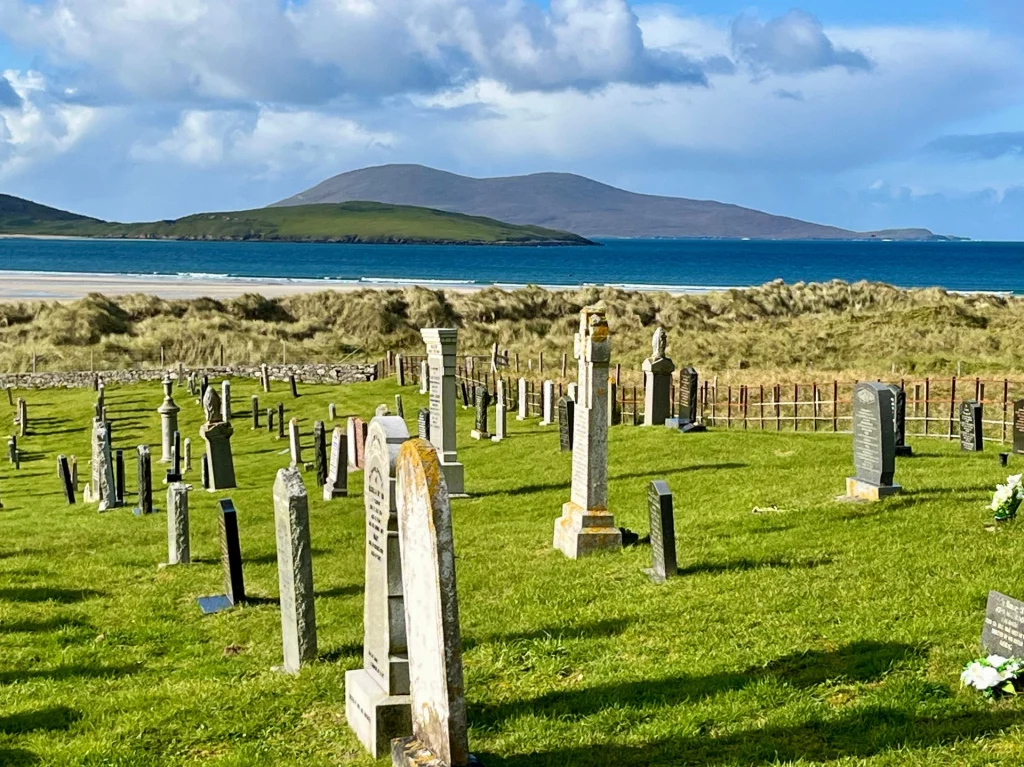 Ein historischer Friedhof mit alten Stein-Grabmalen auf einer saftig grünen Wiese direkt an einer Meeresbucht mit Blick auf entfernte Berge.