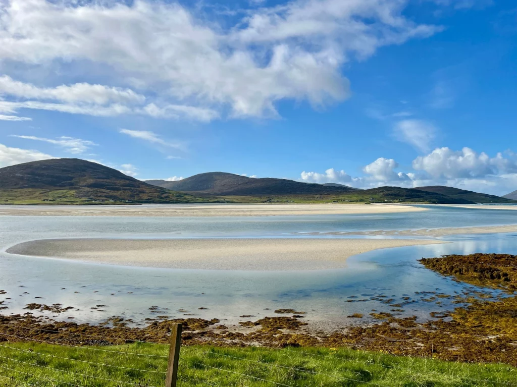Eine weite Küstenlandschaft bei Ebbe mit hellen Sandbänken, ruhigem Wasser und sanften, grünen Hügeln unter einem leicht bewölkten blauen Himmel.