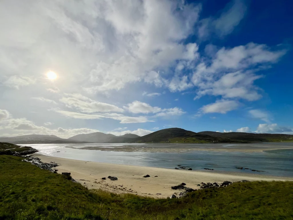 Eine Weitwinkelaufnahme eines hellen Sandstrandes in einer Bucht, umrahmt von dunklen Hügelketten unter einem dramatischen Himmel mit leuchtender Sonne und weißen Wolken.