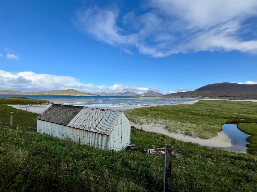 Eine alte, weiß gestrichene Blechhütte mit rostigem Wellblechdach steht auf einer grünen Wiese im Vordergrund. Dahinter erstreckt sich eine weite Bucht mit hellem Sand und flachem Wasser unter einem strahlend blauen Himmel mit feinen Schleierwolken.